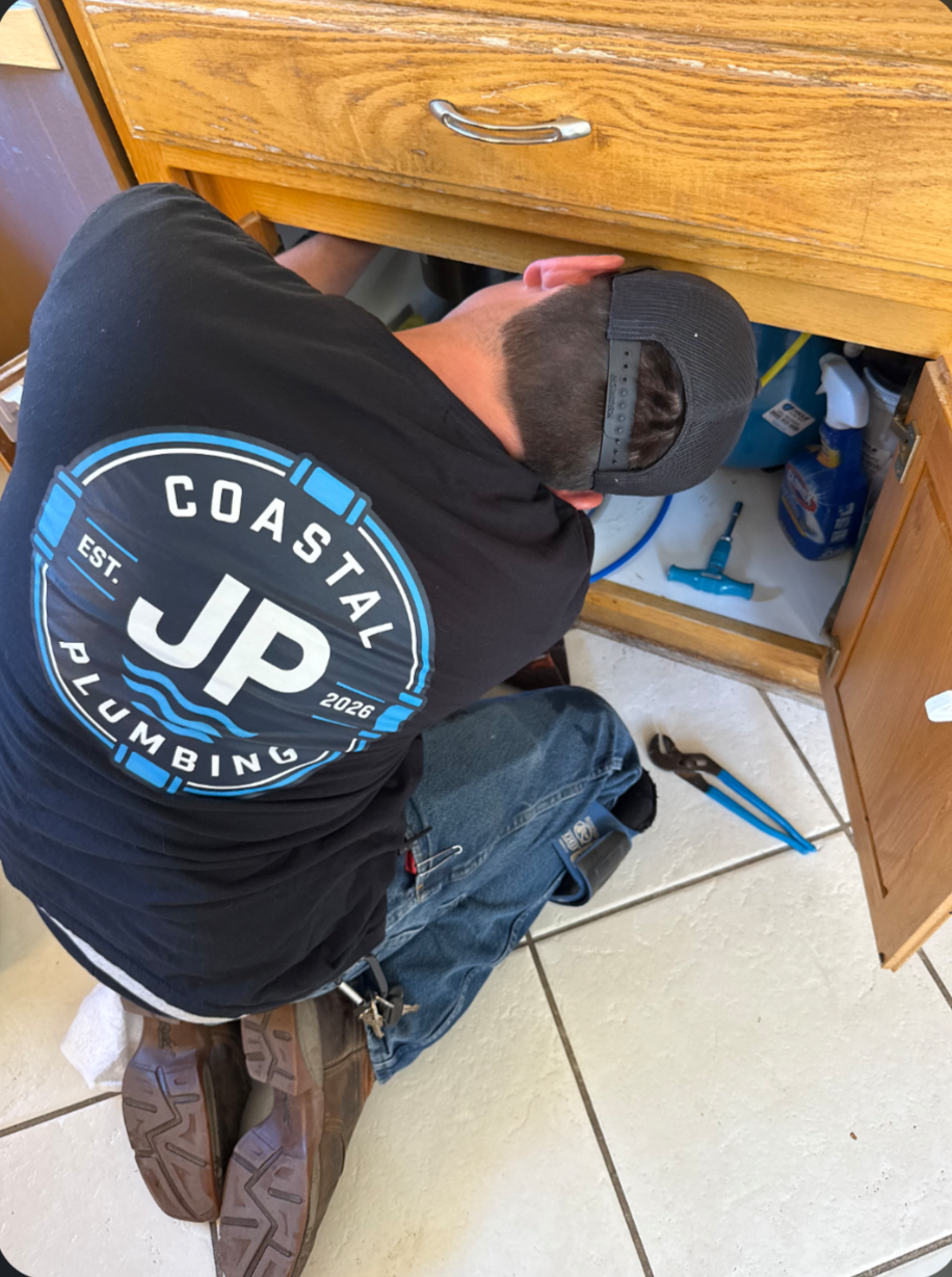 Plumber wearing a JP Coastal Plumbing branded shirt while working inside a kitchen sink cabinet.